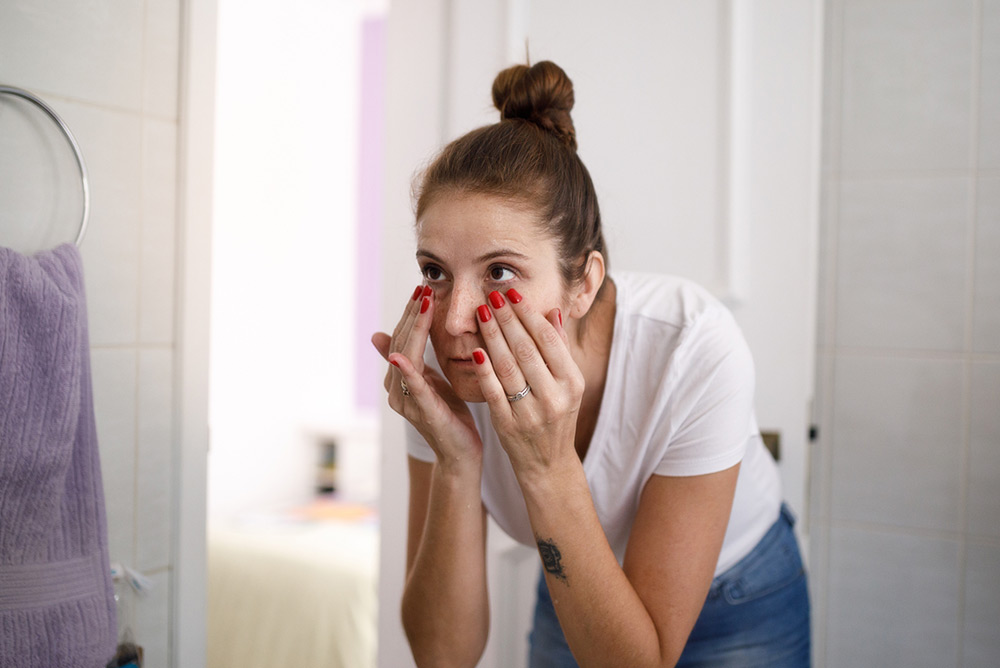 Jeune femme dans salle de bain devant miroir observant des cernes sous les yeux dues à la fatigue