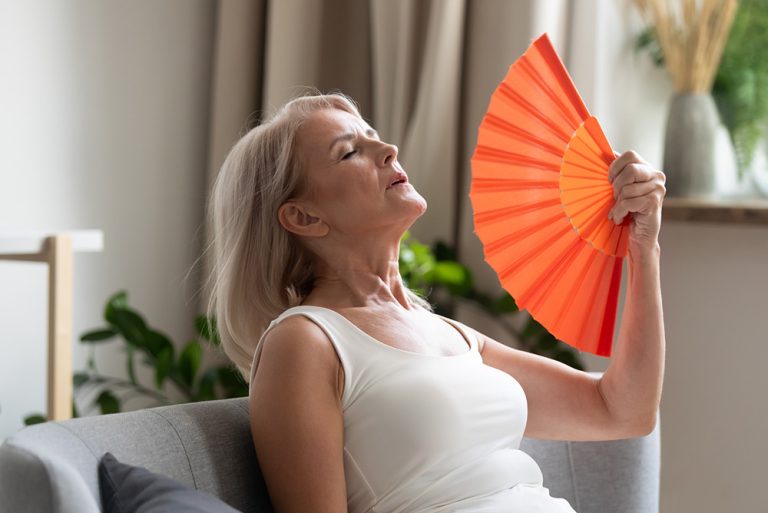 Femme assise sur un canapé ayant des bouffées de chaleur avec un éventail dans la main